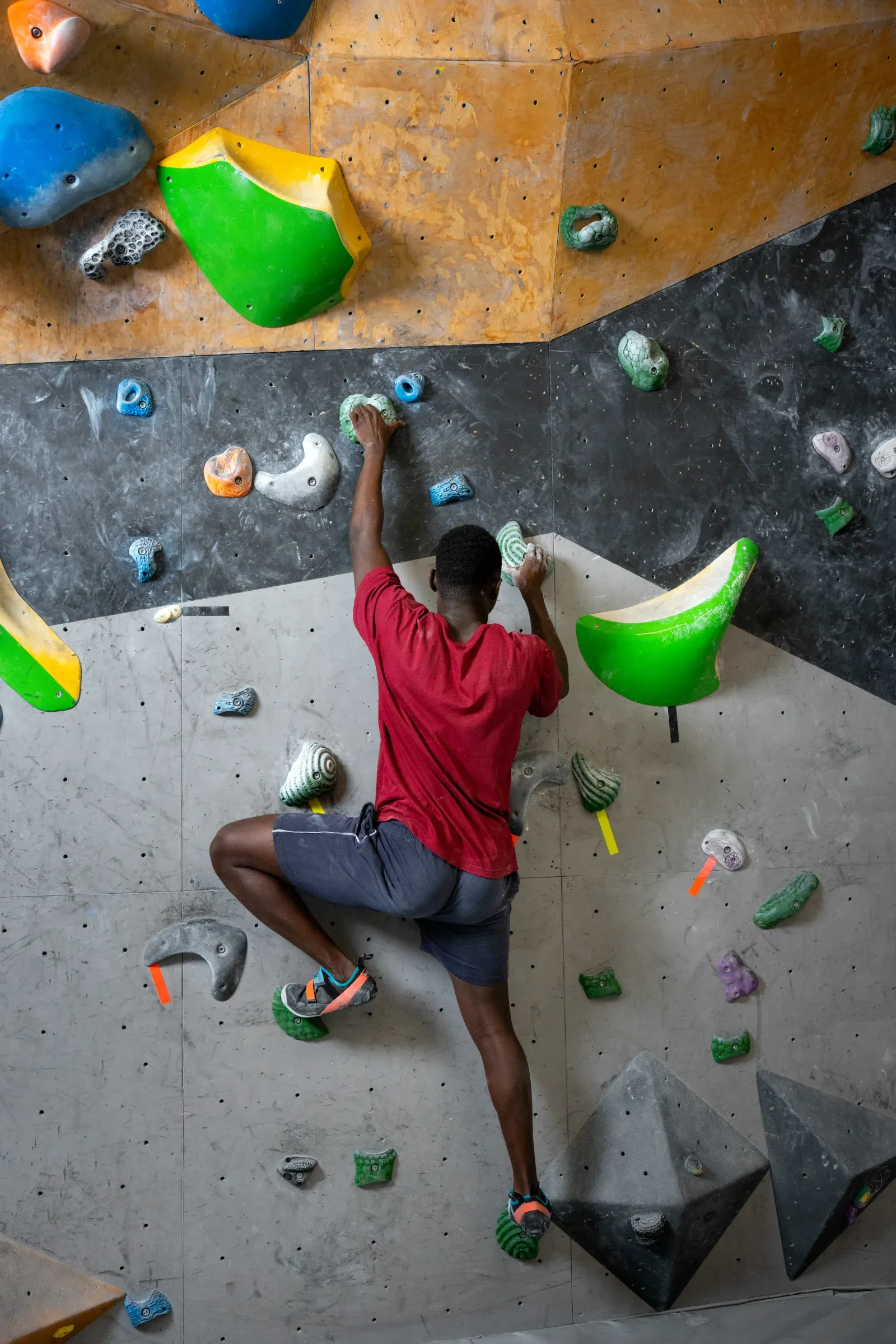 De dos, un grimpeur en tee-shirt rouge progresse sur un mur de bloc, un pied haut sur une prise.